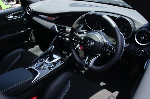 The interior of a modern car featuring a sleek, black leather steering wheel with intricate controls and a prominent logo in the center. The dashboard includes several round dials and a central control panel with a gear stick, buttons, and switches. The design is clean and sophisticated with a focus on ergonomic placement and premium materials.