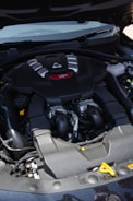 A car engine compartment featuring a prominently labeled V6 engine. The engine has a sleek black cover with silver and red accents. Several hoses and mechanical components are visible around the engine.