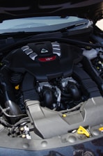 A car engine compartment featuring a prominently labeled V6 engine. The engine has a sleek black cover with silver and red accents. Several hoses and mechanical components are visible around the engine.