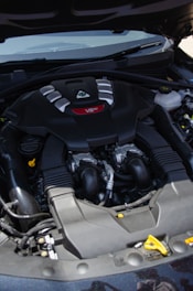A car engine compartment featuring a prominently labeled V6 engine. The engine has a sleek black cover with silver and red accents. Several hoses and mechanical components are visible around the engine.