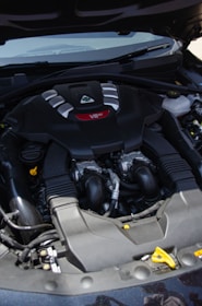 A car engine compartment featuring a prominently labeled V6 engine. The engine has a sleek black cover with silver and red accents. Several hoses and mechanical components are visible around the engine.