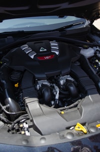 A car engine compartment featuring a prominently labeled V6 engine. The engine has a sleek black cover with silver and red accents. Several hoses and mechanical components are visible around the engine.
