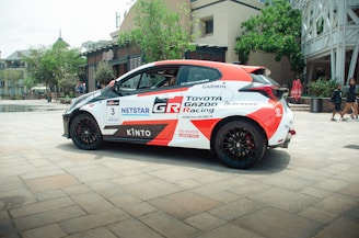 A sleek racing car with a white and red color scheme, prominently displaying logos and decals such as Toyota Gazoo Racing and other sponsors. The car is parked on a paved area, with an urban backdrop including trees, a modern building, and a couple of people walking nearby.
