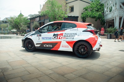 A sleek racing car with a white and red color scheme, prominently displaying logos and decals such as Toyota Gazoo Racing and other sponsors. The car is parked on a paved area, with an urban backdrop including trees, a modern building, and a couple of people walking nearby.