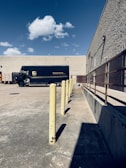 A delivery truck parked in an empty concrete lot near a beige, windowless building. There are yellow bollards aligned in a straight line, and a clear blue sky with a few scattered clouds overhead. To the right is a metal railing along the building's wall.