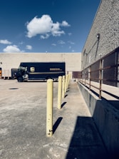 A delivery truck parked in an empty concrete lot near a beige, windowless building. There are yellow bollards aligned in a straight line, and a clear blue sky with a few scattered clouds overhead. To the right is a metal railing along the building's wall.