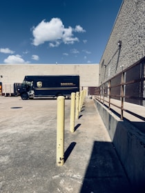 A delivery truck parked in an empty concrete lot near a beige, windowless building. There are yellow bollards aligned in a straight line, and a clear blue sky with a few scattered clouds overhead. To the right is a metal railing along the building's wall.