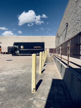 A delivery truck parked in an empty concrete lot near a beige, windowless building. There are yellow bollards aligned in a straight line, and a clear blue sky with a few scattered clouds overhead. To the right is a metal railing along the building's wall.
