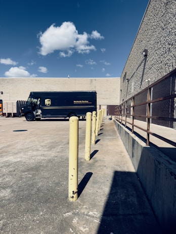 A delivery truck parked in an empty concrete lot near a beige, windowless building. There are yellow bollards aligned in a straight line, and a clear blue sky with a few scattered clouds overhead. To the right is a metal railing along the building's wall.