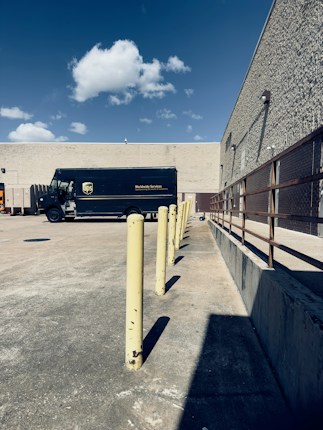 A delivery truck parked in an empty concrete lot near a beige, windowless building. There are yellow bollards aligned in a straight line, and a clear blue sky with a few scattered clouds overhead. To the right is a metal railing along the building's wall.