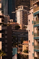 A cityscape featuring a dense arrangement of modern high-rise and mid-rise buildings. Architectural styles include glass exteriors and uniform balconies. Tall trees are interspersed among the structures, adding greenery to the urban setting. A prominently visible sign advertises an event called 'Monaco Auction Week'. The background showcases a mountain, indicating a coastal city.
