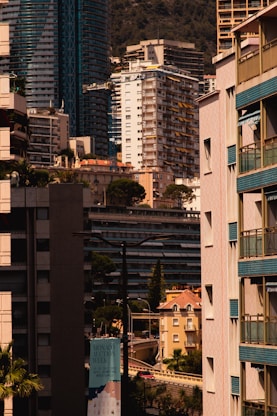 A cityscape featuring a dense arrangement of modern high-rise and mid-rise buildings. Architectural styles include glass exteriors and uniform balconies. Tall trees are interspersed among the structures, adding greenery to the urban setting. A prominently visible sign advertises an event called 'Monaco Auction Week'. The background showcases a mountain, indicating a coastal city.