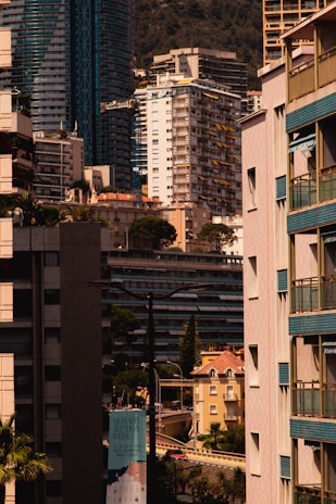 A cityscape featuring a dense arrangement of modern high-rise and mid-rise buildings. Architectural styles include glass exteriors and uniform balconies. Tall trees are interspersed among the structures, adding greenery to the urban setting. A prominently visible sign advertises an event called 'Monaco Auction Week'. The background showcases a mountain, indicating a coastal city.