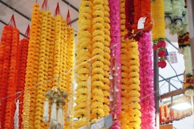 A vibrant display of colorful flower garlands hanging vertically. The garlands are primarily in shades of yellow, orange, and pink. They are arranged densely, creating a lush and festive atmosphere. Some price tags are visible, indicating a market setting. The scene is bright and lively with a focus on the floral arrangements.