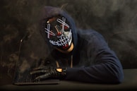 A mysterious figure wearing a hood, illuminated by the glow of a laptop screen displaying classified files.