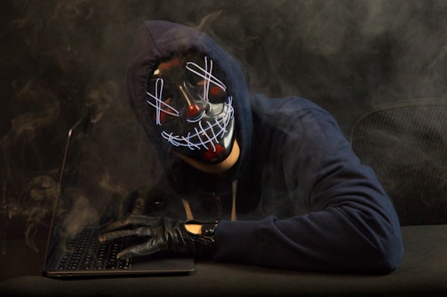 A person wearing a dark hoodie and a distinctive mask with a neon-like grin is sitting at a laptop. Smoke swirls around them, creating a mysterious and dramatic atmosphere. The gloved hand rests on the keyboard, enhancing the sense of secrecy or anonymity.