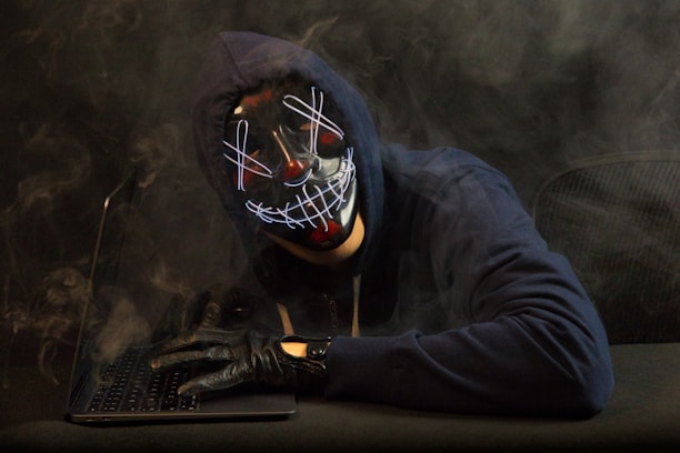 A person wearing a dark hoodie and a distinctive mask with a neon-like grin is sitting at a laptop. Smoke swirls around them, creating a mysterious and dramatic atmosphere. The gloved hand rests on the keyboard, enhancing the sense of secrecy or anonymity.