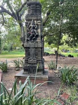A detailed stone sculpture of a deity, possibly Ganesh, stands in a garden. The statue is intricate, with red flowers placed on it as offerings. Surrounding the structure are lush green trees and plants, creating a serene and natural environment.