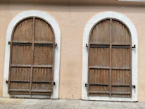 There are two arched wooden doors with a weathered appearance set in a light beige wall. The doors are secured with black iron hinges.