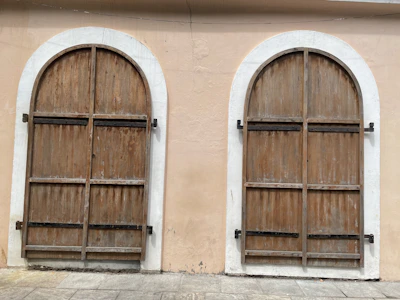 There are two arched wooden doors with a weathered appearance set in a light beige wall. The doors are secured with black iron hinges.