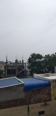 A partially constructed building with exposed rebar in an urban area. There are several trees nearby, adding greenery to the scene. Adjacent buildings have corrugated blue roofs and weathered brick exterior walls. The sky appears overcast and somewhat subdued.