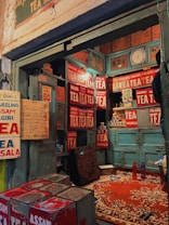 A rustic tea shop interior with vintage signs advertising various types of tea like Assam, Nilgiri, and Kashmiri. The walls are adorned with numerous red and white tea signs, and there is a collection of tea tins and cans. A worn-out rug covers part of the floor, and old wooden cabinets are visible along with a clock and some miscellaneous items.
