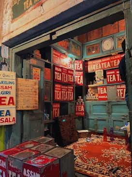A rustic tea shop interior with vintage signs advertising various types of tea like Assam, Nilgiri, and Kashmiri. The walls are adorned with numerous red and white tea signs, and there is a collection of tea tins and cans. A worn-out rug covers part of the floor, and old wooden cabinets are visible along with a clock and some miscellaneous items.