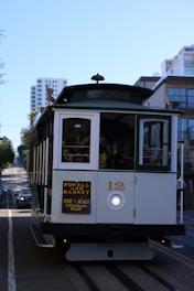 A traditional San Francisco cable car is moving along a hilly street surrounded by modern buildings. Passengers can be seen seated inside, enjoying the ride. The sky is clear and blue, suggesting a sunny day.