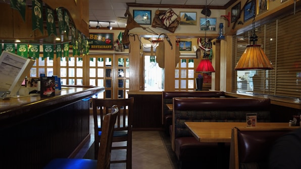 A cozy restaurant interior featuring wooden booths and tables with a combination of warm and muted lighting. The decor includes colorful pendant lamps, nautical-themed items like a ship's wheel, and various framed pictures on the walls. Green banners hang from the ceiling, and a counter area is visible on the left.