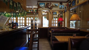 A cozy restaurant interior featuring wooden booths and tables with a combination of warm and muted lighting. The decor includes colorful pendant lamps, nautical-themed items like a ship's wheel, and various framed pictures on the walls. Green banners hang from the ceiling, and a counter area is visible on the left.