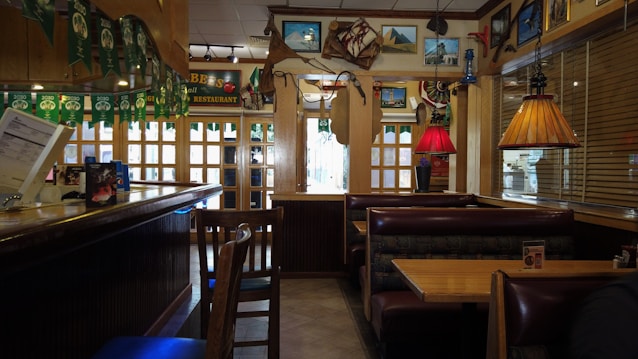 A cozy restaurant interior featuring wooden booths and tables with a combination of warm and muted lighting. The decor includes colorful pendant lamps, nautical-themed items like a ship's wheel, and various framed pictures on the walls. Green banners hang from the ceiling, and a counter area is visible on the left.
