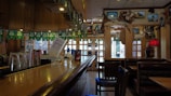 A dimly lit, cozy bar with a wooden countertop and glassware on display. Wall decorations include framed pictures, maps, and various ornaments. Green party banners hang above, and seating areas with wooden chairs and leather booths are visible.