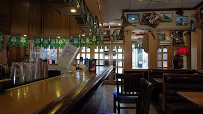 A dimly lit, cozy bar with a wooden countertop and glassware on display. Wall decorations include framed pictures, maps, and various ornaments. Green party banners hang above, and seating areas with wooden chairs and leather booths are visible.