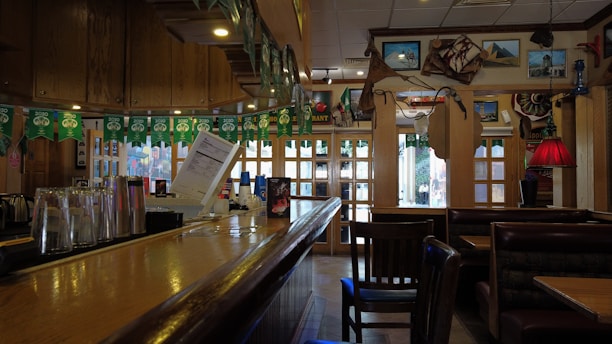 A dimly lit, cozy bar with a wooden countertop and glassware on display. Wall decorations include framed pictures, maps, and various ornaments. Green party banners hang above, and seating areas with wooden chairs and leather booths are visible.