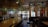 A dimly lit, cozy bar with a wooden countertop and glassware on display. Wall decorations include framed pictures, maps, and various ornaments. Green party banners hang above, and seating areas with wooden chairs and leather booths are visible.