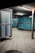 A dimly lit industrial corridor features blue paneling and metallic structures. The floor is tiled with large stone slabs, adding a sense of geometric order. Fluorescent lights cast a stark illumination from the ceiling. A person, only partially visible, appears to be walking around the corner.