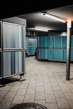 A dimly lit industrial corridor features blue paneling and metallic structures. The floor is tiled with large stone slabs, adding a sense of geometric order. Fluorescent lights cast a stark illumination from the ceiling. A person, only partially visible, appears to be walking around the corner.
