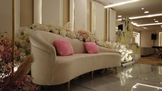 A luxurious sofa adorned with pink cushions is surrounded by a lavish arrangement of white and pink flowers. The setting features elegant lighting with ceiling fixtures, creating a sophisticated atmosphere. The background includes a decorated panel and an electronic keyboard, suggesting an event space.