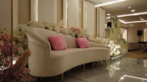 A luxurious sofa adorned with pink cushions is surrounded by a lavish arrangement of white and pink flowers. The setting features elegant lighting with ceiling fixtures, creating a sophisticated atmosphere. The background includes a decorated panel and an electronic keyboard, suggesting an event space.