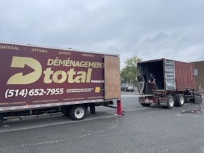 A moving truck with company branding is parked next to an open shipping container on a trailer. Two people are handling items near the container, while a car is parked in the background. The sky is overcast, and the ground appears to be asphalt.