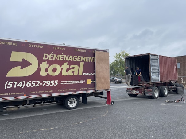 A moving truck with company branding is parked next to an open shipping container on a trailer. Two people are handling items near the container, while a car is parked in the background. The sky is overcast, and the ground appears to be asphalt.