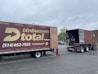 A moving truck with company branding is parked next to an open shipping container on a trailer. Two people are handling items near the container, while a car is parked in the background. The sky is overcast, and the ground appears to be asphalt.