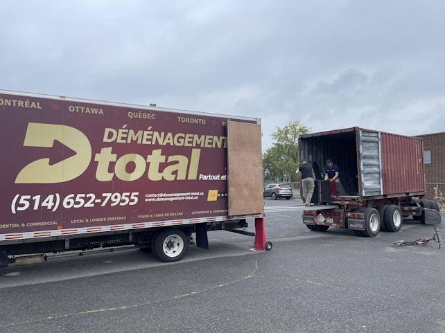 A moving truck with company branding is parked next to an open shipping container on a trailer. Two people are handling items near the container, while a car is parked in the background. The sky is overcast, and the ground appears to be asphalt.