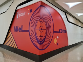 A large wall with an orange background features a graphic design centered around a circular stadium illustration. The words 'WeL' and 'come' are prominently displayed, with a gap presumably intended for the missing letters 'c' and 'o'. The design incorporates elements like diamonds and a football field, with text indicating 'FIFA World Cup Qatar 2022'. The wall has a modern design with a black border and is part of an indoor space with shiny flooring.