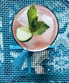 A refreshing pink beverage in a glass, topped with ice cubes, a slice of cucumber, and sprigs of mint. The drink is placed on a vibrant blue mesh surface that adds a textured background.