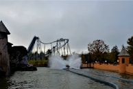 A theme park scene featuring a roller coaster with a water splash ride. The roller coaster is a large structure with paths that twist and turn, located in the background. In the foreground, there is a splash zone where a ride generates a large spray of water upon impact. Trees and park buildings with rustic designs surround the area, adding a natural and adventurous atmosphere.