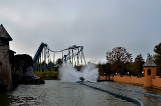 A theme park scene featuring a roller coaster with a water splash ride. The roller coaster is a large structure with paths that twist and turn, located in the background. In the foreground, there is a splash zone where a ride generates a large spray of water upon impact. Trees and park buildings with rustic designs surround the area, adding a natural and adventurous atmosphere.