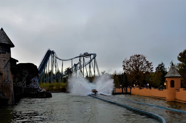 A theme park scene featuring a roller coaster with a water splash ride. The roller coaster is a large structure with paths that twist and turn, located in the background. In the foreground, there is a splash zone where a ride generates a large spray of water upon impact. Trees and park buildings with rustic designs surround the area, adding a natural and adventurous atmosphere.