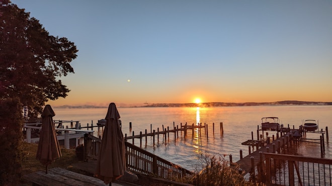 A serene lakeside scene at sunrise with the sun low on the horizon, creating a warm glow on the water. Wooden docks extend into the lake, flanked by boats, and trees frame the scene on the left. Closed umbrellas stand on the deck area, and mist hovers just above the water's surface.