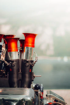 A mechanical component with several cylindrical pipes extending vertically, each topped with a flared red end. The structure appears industrial, with shiny metallic surfaces and visible bolts and screws. The background is blurred, suggesting an outdoor setting with greenery.